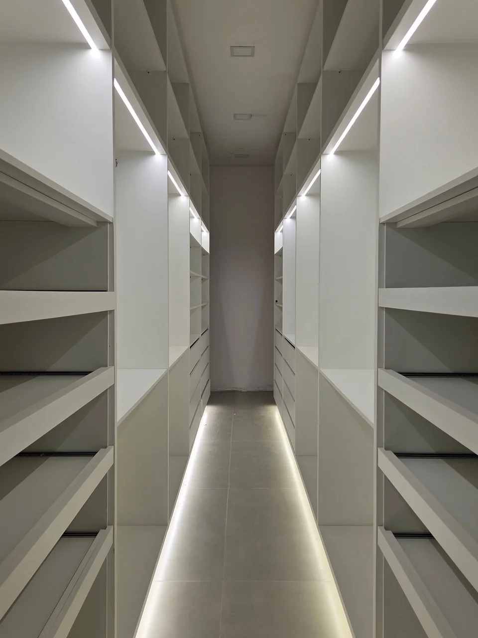 Closet com prateleiras abertas, gavetas e iluminação linear no teto e na base dos armários.